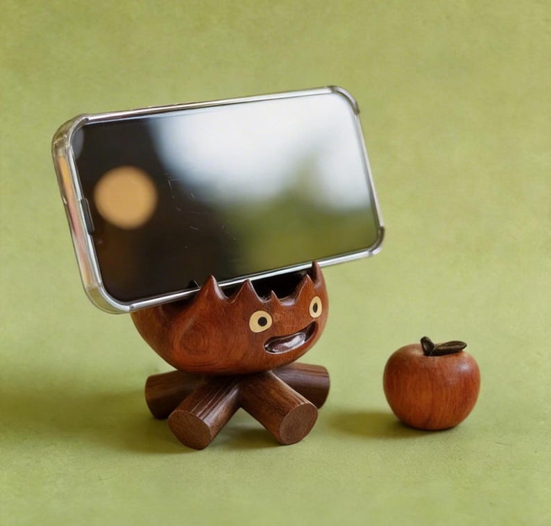 May include: A wooden phone stand shaped like a cartoon character with a smiling face and a small wooden apple. The stand is holding a phone with a clear case. The stand and apple are brown, set against a green background.