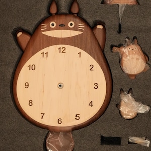May include: A wooden wall clock shaped like a cartoon creature with a dark brown body, light wood face, and black numerals from 1 to 12. The clock includes separate wooden components, including a small umbrella and two smaller figures, all packaged in foam.