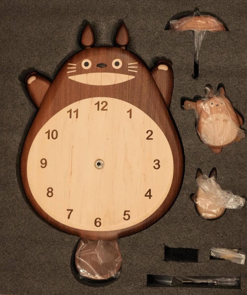 May include: A wooden wall clock shaped like a cartoon creature with a dark brown body, light wood face, and black numerals from 1 to 12. The clock includes separate wooden components, including a small umbrella and two smaller figures, all packaged in foam.