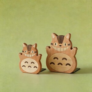 May include: Two wooden figurines of a smiling cat-like creature with a light brown body and dark brown accents. The figurines have a friendly expression and are standing on a black base. The background is a solid light green.