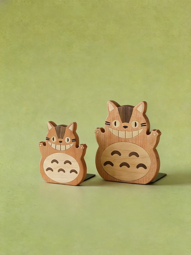 May include: Two wooden figurines of a smiling cat-like creature with a light brown body and dark brown accents. The figurines have a friendly expression and are standing on a black base. The background is a solid light green.