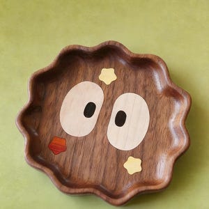 May include: A decorative wooden tray with a whimsical design. The tray is a dark brown colour with a wavy edge. It features inlaid light wood and black details to create a face with large eyes, a mouth, and star accents. The background is a solid green.