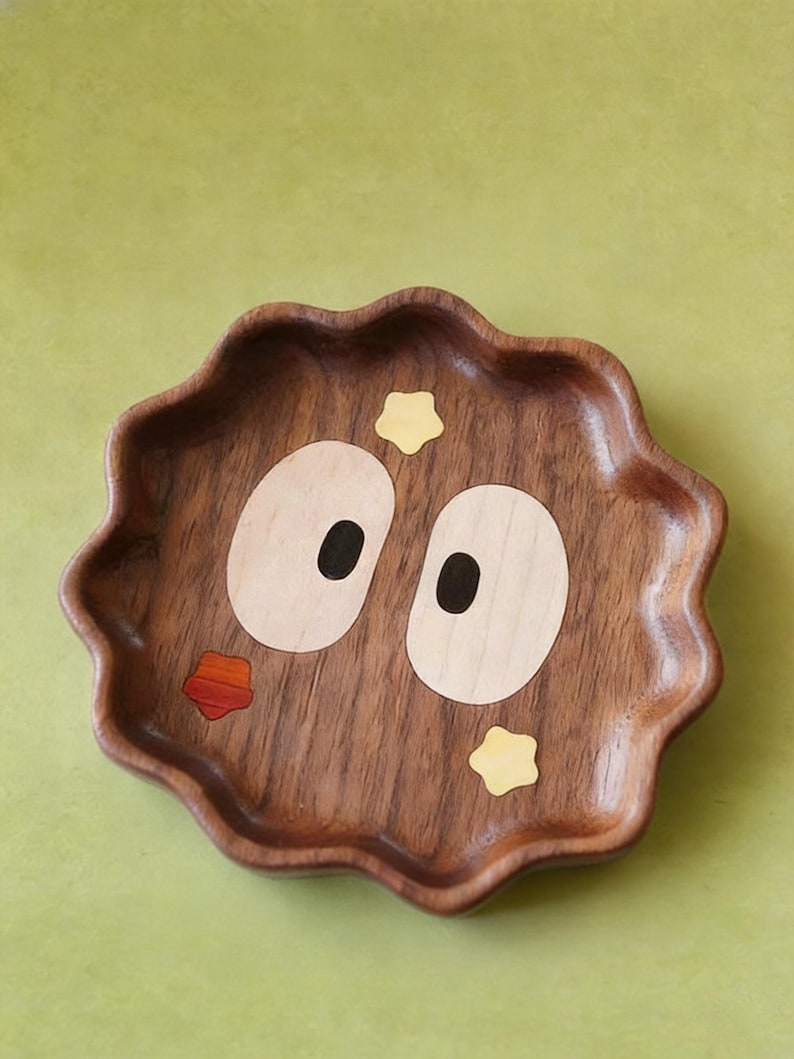 May include: A decorative wooden tray with a whimsical design. The tray is a dark brown colour with a wavy edge. It features inlaid light wood and black details to create a face with large eyes, a mouth, and star accents. The background is a solid green.