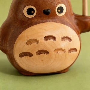 Puede incluir: Figura de madera de un personaje de dibujos animados con cuerpo redondo, ojos grandes y nariz peque&ntilde;a negra. La figura es principalmente marr&oacute;n con vientre de madera m&aacute;s clara y cortes decorativos. Un acento verde es visible en la parte superior.