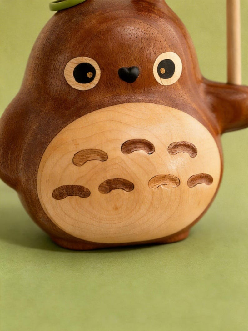 Puede incluir: Figura de madera de un personaje de dibujos animados con cuerpo redondo, ojos grandes y nariz peque&ntilde;a negra. La figura es principalmente marr&oacute;n con vientre de madera m&aacute;s clara y cortes decorativos. Un acento verde es visible en la parte superior.