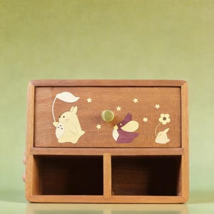 K&ouml;nnte beinhalten: Ein h&ouml;lzerner Schreibtisch-Organizer mit einer Schublade und zwei offenen F&auml;chern. Die Schublade ist mit Zeichentrickfiguren und Sternen verziert. Der Organizer hat einen Messingknopf und ist aus hellbraunem Holz gefertigt.