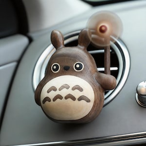 May include: A wooden car air vent clip featuring a cartoon character design. The brown figure has large eyes, a white belly, and a small fan. The fan is attached to a wooden stick and spins. The clip is attached to a car vent.