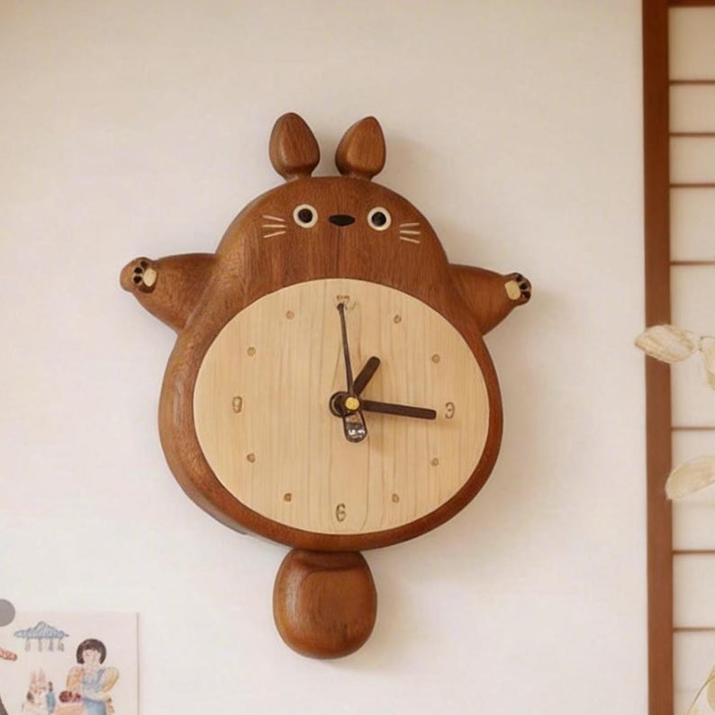 May include: A wooden wall clock shaped like a cartoon creature with a dark brown body, two ears, and a light beige circular face. The clock features black hands, small paw details on the outstretched arms, and a rounded tail hanging at the bottom.