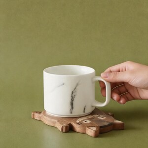 May include: A white ceramic mug with a black marble design sits on a wooden coaster shaped like a cat. The coaster is brown with carved details. The mug has a handle and is held by a person's hand. The background is a solid olive green.