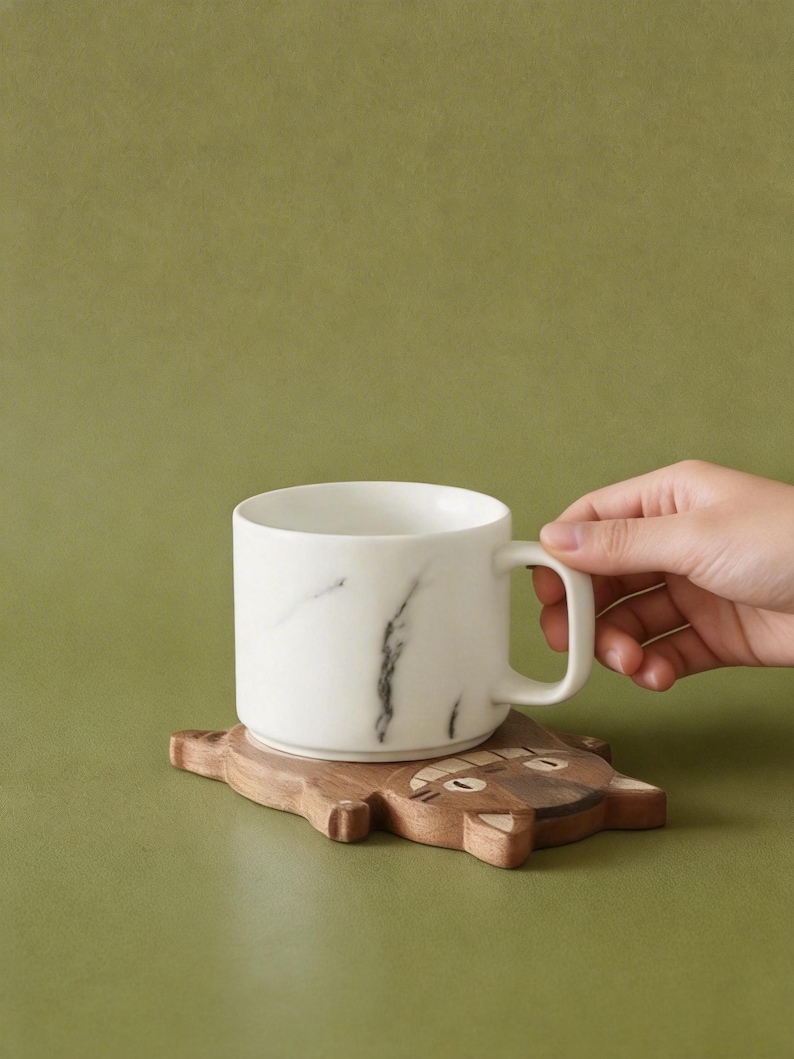 May include: A white ceramic mug with a black marble design sits on a wooden coaster shaped like a cat. The coaster is brown with carved details. The mug has a handle and is held by a person's hand. The background is a solid olive green.