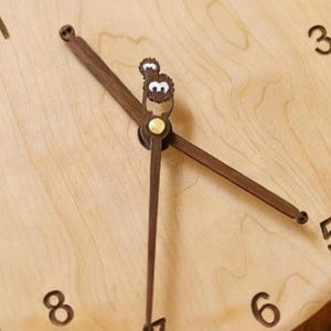 May include: A light-coloured wooden wall clock face featuring dark brown wooden hands. The hour hand is shaped like a small creature with white eyes, while the minute hand is a simple dark brown pointer. Black numerals 3, 6, and 8 are visible.