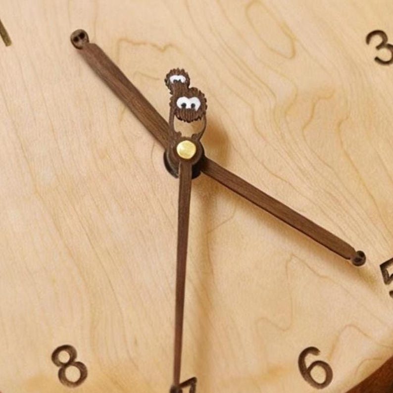 May include: A light-coloured wooden wall clock face featuring dark brown wooden hands. The hour hand is shaped like a small creature with white eyes, while the minute hand is a simple dark brown pointer. Black numerals 3, 6, and 8 are visible.