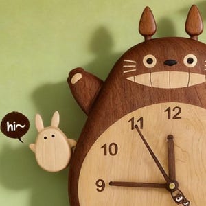 May include: A wooden wall clock shaped like a large brown creature with pointed ears, a wide smiling mouth, and a light-coloured belly featuring black numbers. A small light-coloured figure stands to the left with a speech bubble containing the text hi~.