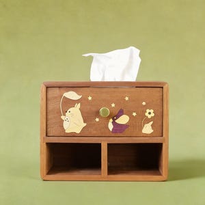 K&ouml;nnte beinhalten: Holz-Taschentuchbox mit einer Schublade und zwei unteren F&auml;chern. Die Box ist mit Intarsien aus Holz mit einem Hasen, der ein Blatt h&auml;lt, einem lila-wei&szlig;en Vogel und einer Blume verziert. Ein wei&szlig;es Taschentuch ragt aus der Oberseite heraus.