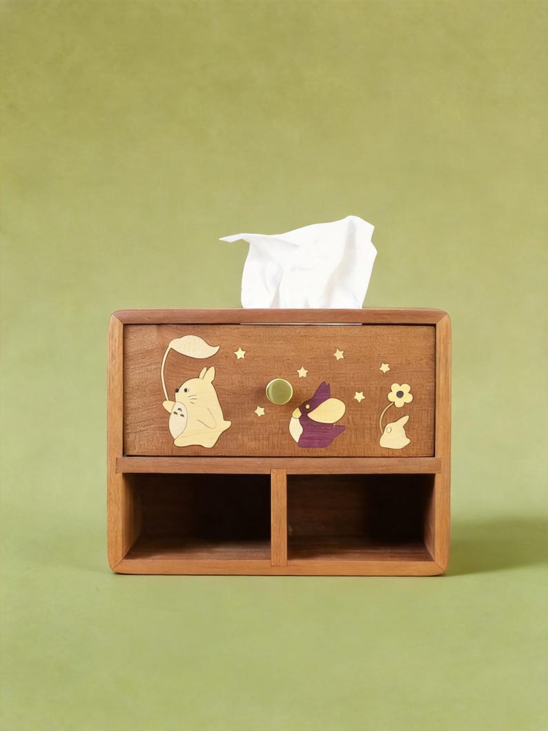 K&ouml;nnte beinhalten: Holz-Taschentuchbox mit einer Schublade und zwei unteren F&auml;chern. Die Box ist mit Intarsien aus Holz mit einem Hasen, der ein Blatt h&auml;lt, einem lila-wei&szlig;en Vogel und einer Blume verziert. Ein wei&szlig;es Taschentuch ragt aus der Oberseite heraus.