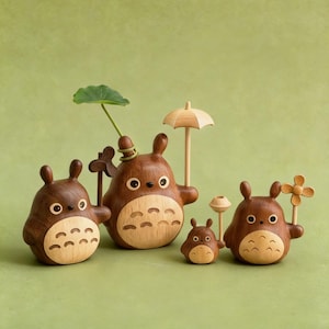 Puede incluir: Colecci&oacute;n de cinco figuras de madera de Totoro, cada una con un cuerpo marr&oacute;n y una barriga de madera clara. Algunas sostienen accesorios como un paraguas, una flor y una hoja. Las figuras est&aacute;n sobre un fondo verde.