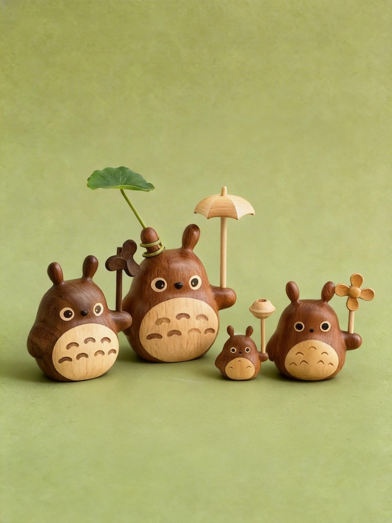 Puede incluir: Colecci&oacute;n de cinco figuras de madera de Totoro, cada una con un cuerpo marr&oacute;n y una barriga de madera clara. Algunas sostienen accesorios como un paraguas, una flor y una hoja. Las figuras est&aacute;n sobre un fondo verde.