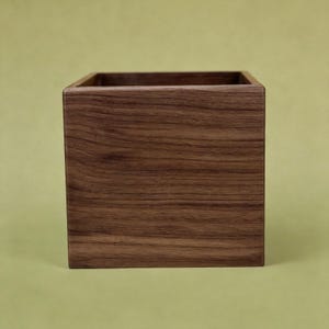 May include: A square wooden box with a rich brown finish. The box has a smooth, polished surface and a simple, minimalist design. The open top reveals the hollow interior, perfect for storage or display.