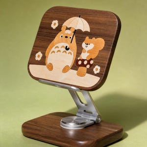 May include: A wooden phone stand featuring a dark walnut base and a rectangular support panel with a light wood inlay. The inlay depicts a cartoon creature holding an umbrella next to a squirrel on a mushroom, surrounded by small flower shapes.