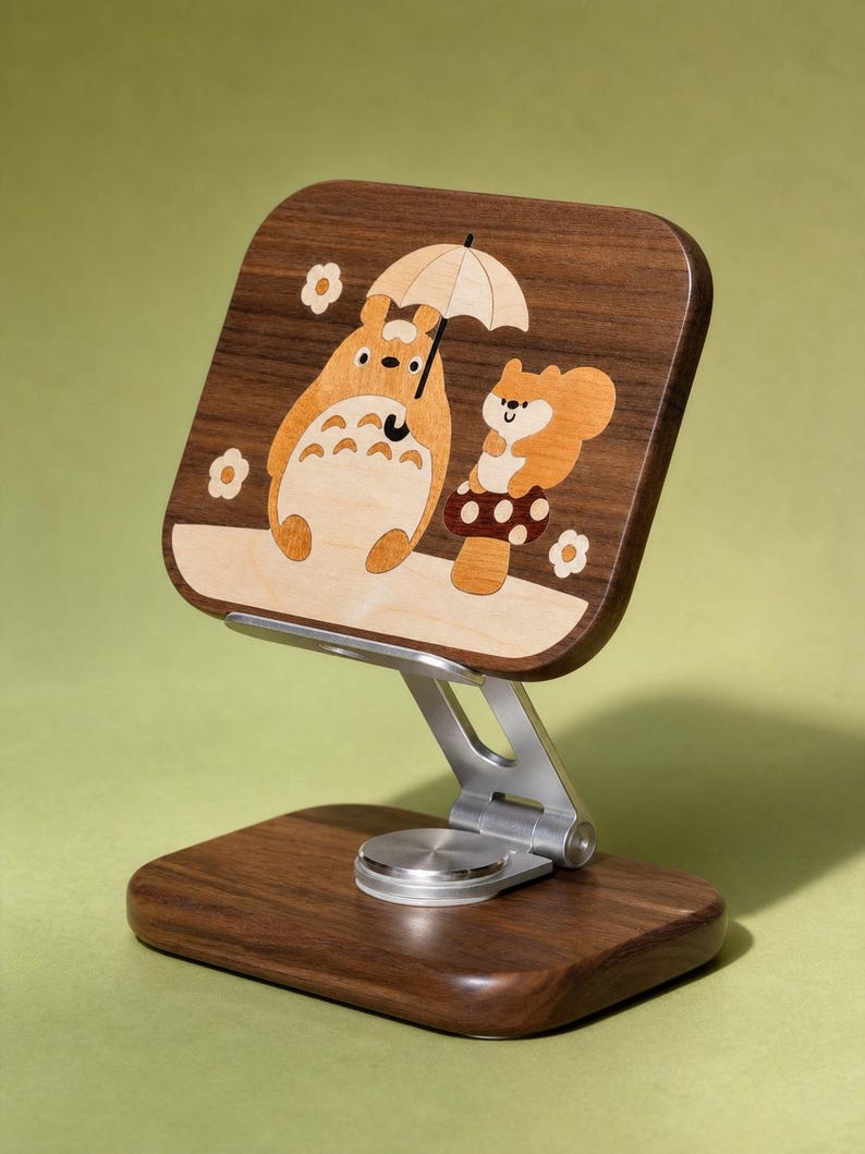 May include: A wooden phone stand featuring a dark walnut base and a rectangular support panel with a light wood inlay. The inlay depicts a cartoon creature holding an umbrella next to a squirrel on a mushroom, surrounded by small flower shapes.