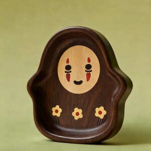 May include: A wooden decorative tray featuring a character design with a light brown face, black eyes, and red accents. The tray is dark brown with three yellow and red flower designs.