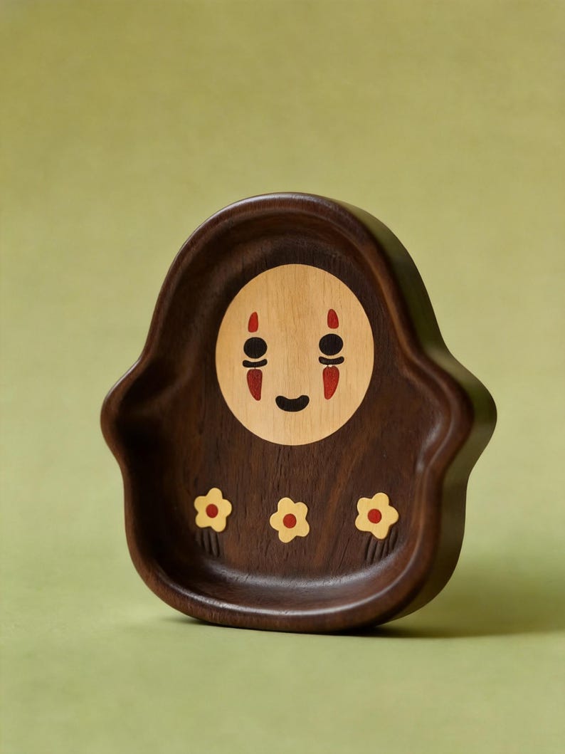 May include: A wooden decorative tray featuring a character design with a light brown face, black eyes, and red accents. The tray is dark brown with three yellow and red flower designs.