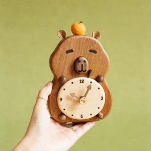 以下が含まれることがあります： 木製のクマ型時計。明るい木製の文字盤が付いています。時計の頭には小さなオレンジ色の果物があります。時計の針は2時10分を指しています。時計は緑色の背景に対して手で持たれています。
