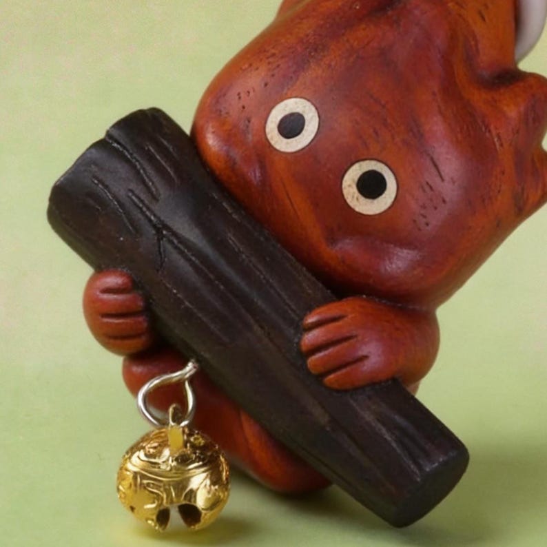 May include: A handcrafted wooden figurine of a creature with a reddish-brown body, holding a dark brown log. The figurine has large, round eyes and a small, gold-colored bell hanging from its hand. The bell has intricate carvings.