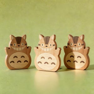 May include: Three wooden figurines of a smiling cartoon cat with a light brown body and dark brown accents. The cat has a large grin and a series of curved shapes on its belly. The figurines are arranged in a row against a green background.