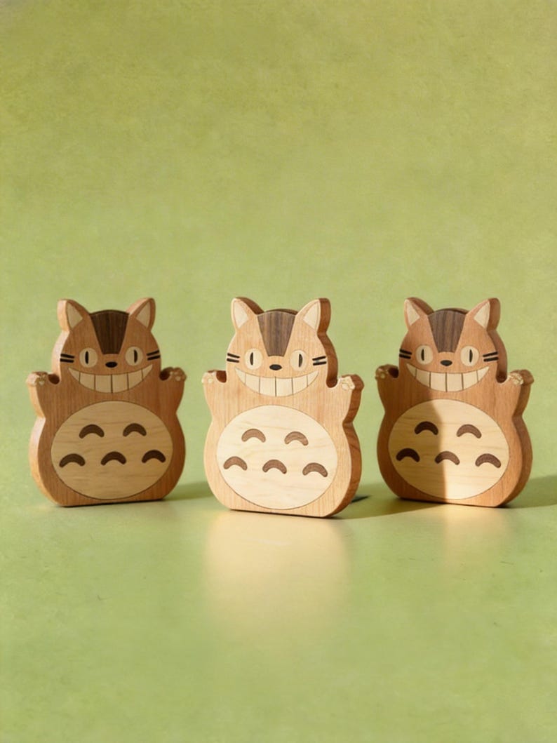 May include: Three wooden figurines of a smiling cartoon cat with a light brown body and dark brown accents. The cat has a large grin and a series of curved shapes on its belly. The figurines are arranged in a row against a green background.