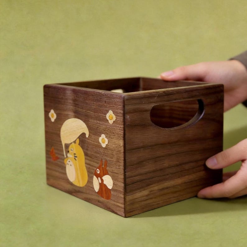 May include: A wooden storage box with a dark brown finish. The box features a cut-out handle and is decorated with inlaid wood designs of a squirrel holding an umbrella and a small rabbit. The box is held by a person.