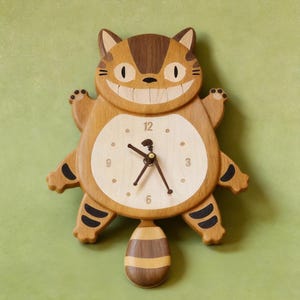 Puede incluir: Reloj de pared de madera con forma de gato de dibujos animados sonriente. El reloj tiene una esfera de color claro con números y manecillas negras. El cuerpo del gato es marrón claro con rayas y detalles en marrón oscuro. El reloj mide unos 30 cm de altura.