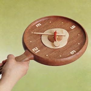 Könnte beinhalten: Eine runde Wanduhr aus Holz in Form einer Bratpfanne mit Griff. Das Zifferblatt hat einen hellen Holzkreis in der Mitte, einen Stundenzeiger in Form einer roten Chilischote, einen Minutenzeiger als Löffel und weiße Ziffern an den Positionen 3, 6, 9 und 12.