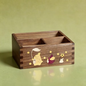 May include: A rectangular wooden desk organiser with three compartments. The front panel features inlaid wood designs of a yellow creature holding a leaf, a purple bird-like figure, a small white ghost, a yellow flower, and several small yellow stars.