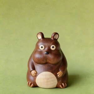 May include: A handcrafted wooden figurine of a round, brown animal with small ears, round eyes, and a light-colored belly. The figurine has a smooth finish and is set against a green background.