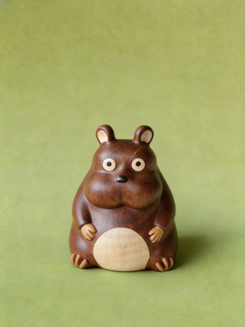 May include: A handcrafted wooden figurine of a round, brown animal with small ears, round eyes, and a light-colored belly. The figurine has a smooth finish and is set against a green background.