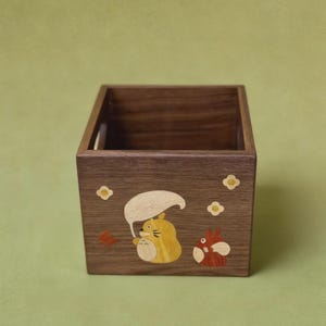 May include: A square wooden box with a dark brown finish. The box is decorated with inlaid cartoon characters in yellow, orange, and white. The background is a solid light green.