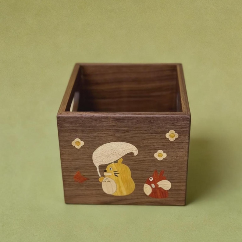 May include: A square wooden box with a dark brown finish. The box is decorated with inlaid cartoon characters in yellow, orange, and white. The background is a solid light green.