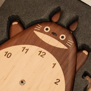 May include: A dark brown wooden wall clock shaped like a cartoon creature with two ears, a light beige circular centre, and dark brown numbers from one to twelve. The creature has a wide, light beige mouth, black eyes, and whiskers.