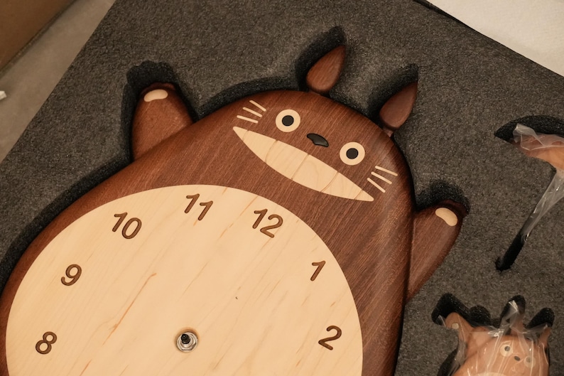 May include: A dark brown wooden wall clock shaped like a cartoon creature with two ears, a light beige circular centre, and dark brown numbers from one to twelve. The creature has a wide, light beige mouth, black eyes, and whiskers.