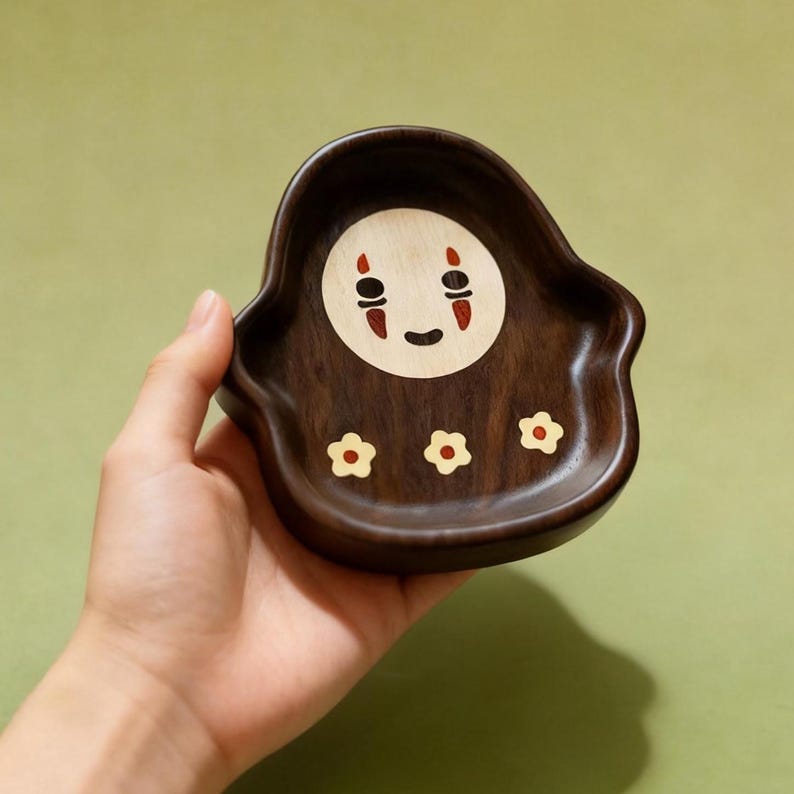 May include: A dark brown wooden tray with a No-Face design. The tray has a rounded shape with a circular face in the center, featuring red and black details. Three small flower inlays are at the bottom. The tray is held by a hand.