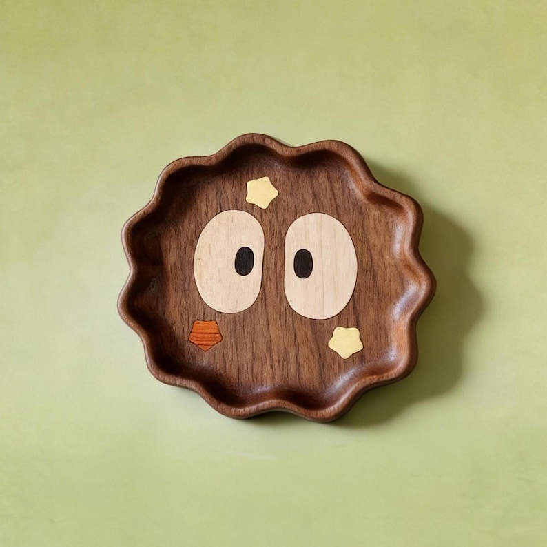 May include: A decorative wooden tray with a cartoon face design. The brown tray has a wavy edge and inlaid light wood and dark wood details to create eyes, a beak, and star accents. A charming piece for home decor.