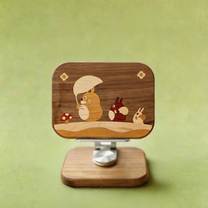 May include: A rectangular wooden phone stand featuring a dark walnut base and a matching upright panel. The panel displays an inlaid wood scene of three cartoon creatures, one holding an umbrella, next to a small red mushroom with white spots.