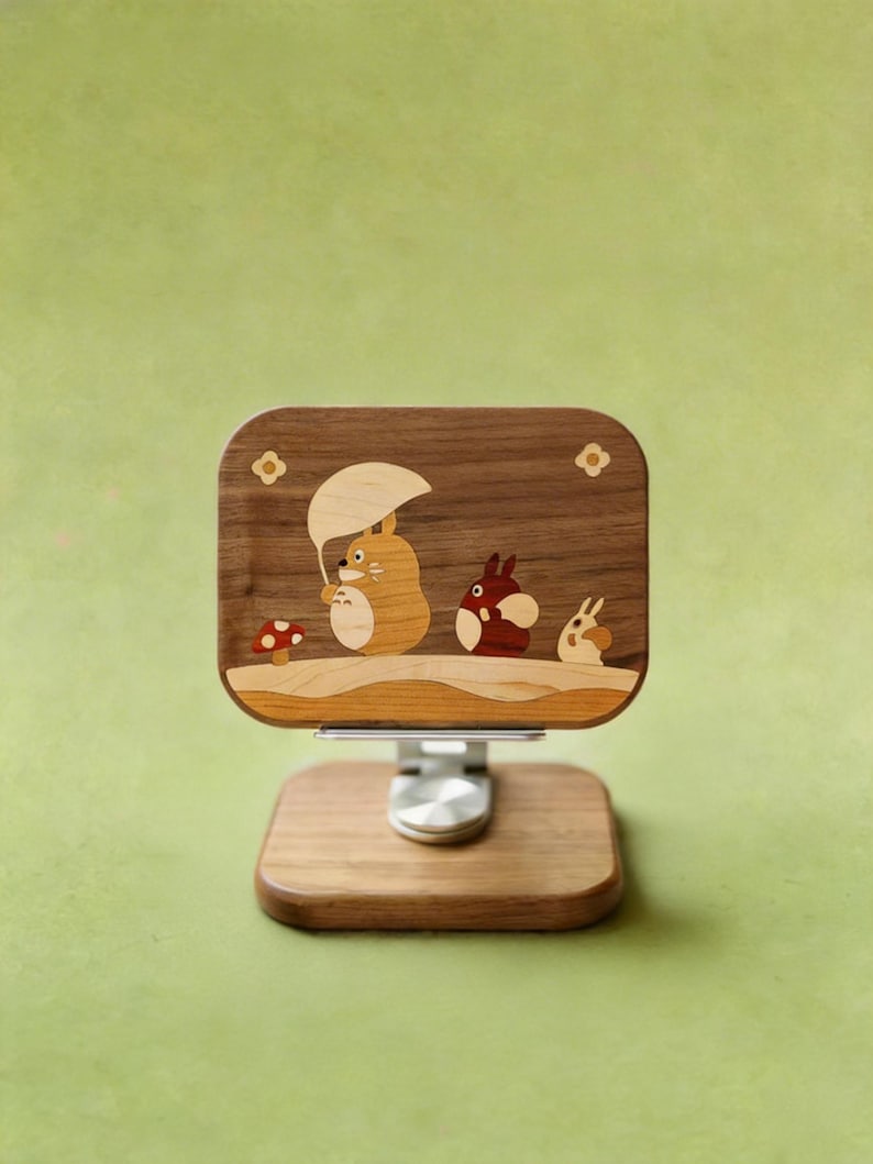 May include: A rectangular wooden phone stand featuring a dark walnut base and a matching upright panel. The panel displays an inlaid wood scene of three cartoon creatures, one holding an umbrella, next to a small red mushroom with white spots.
