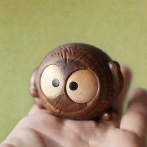 Puede incluir: Peque&ntilde;a figura de b&uacute;ho de madera con ojos grandes y redondos y una textura tallada detallada. El b&uacute;ho es de color marr&oacute;n c&aacute;lido con ojos de color beige claro y pupilas negras. La figura se sostiene en una mano sobre un fondo verde suave.