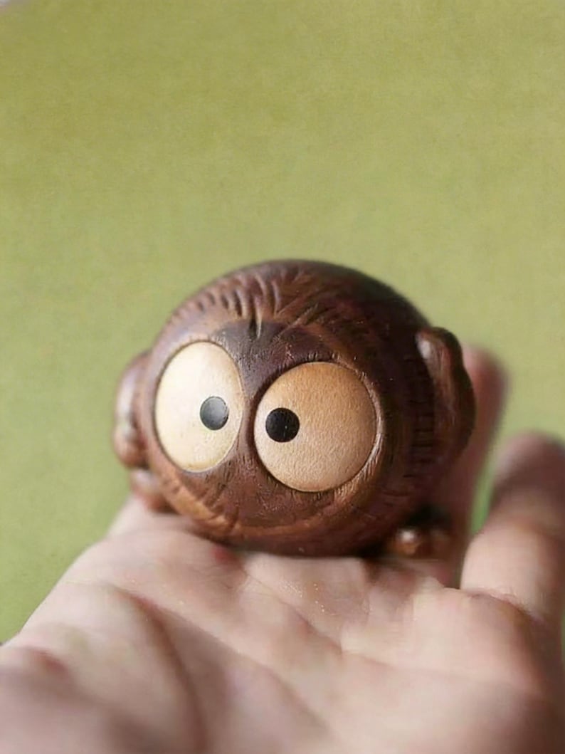 Puede incluir: Peque&ntilde;a figura de b&uacute;ho de madera con ojos grandes y redondos y una textura tallada detallada. El b&uacute;ho es de color marr&oacute;n c&aacute;lido con ojos de color beige claro y pupilas negras. La figura se sostiene en una mano sobre un fondo verde suave.