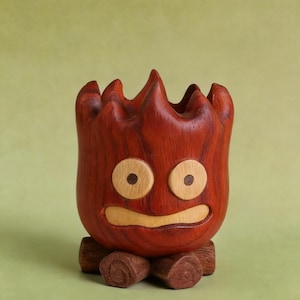 May include: A handcrafted wooden planter shaped like a cartoon character with a fiery appearance. The planter is primarily reddish-brown with lighter wood accents for the eyes and mouth. It sits on a base made of small wooden logs.