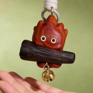 May include: A wooden keychain featuring a stylized, cartoonish flame character with a dark brown log base. The flame is reddish-brown with two white-rimmed eyes. A small gold-colored bell hangs below. The keychain is attached to a white cord.