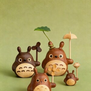 Puede incluir: Una colecci&oacute;n de figuras de madera de Totoro de varios tama&ntilde;os. Las figuras son marrones con vientres de madera clara y presentan peque&ntilde;os accesorios como una hoja, un paraguas y un abanico. El fondo es verde liso.