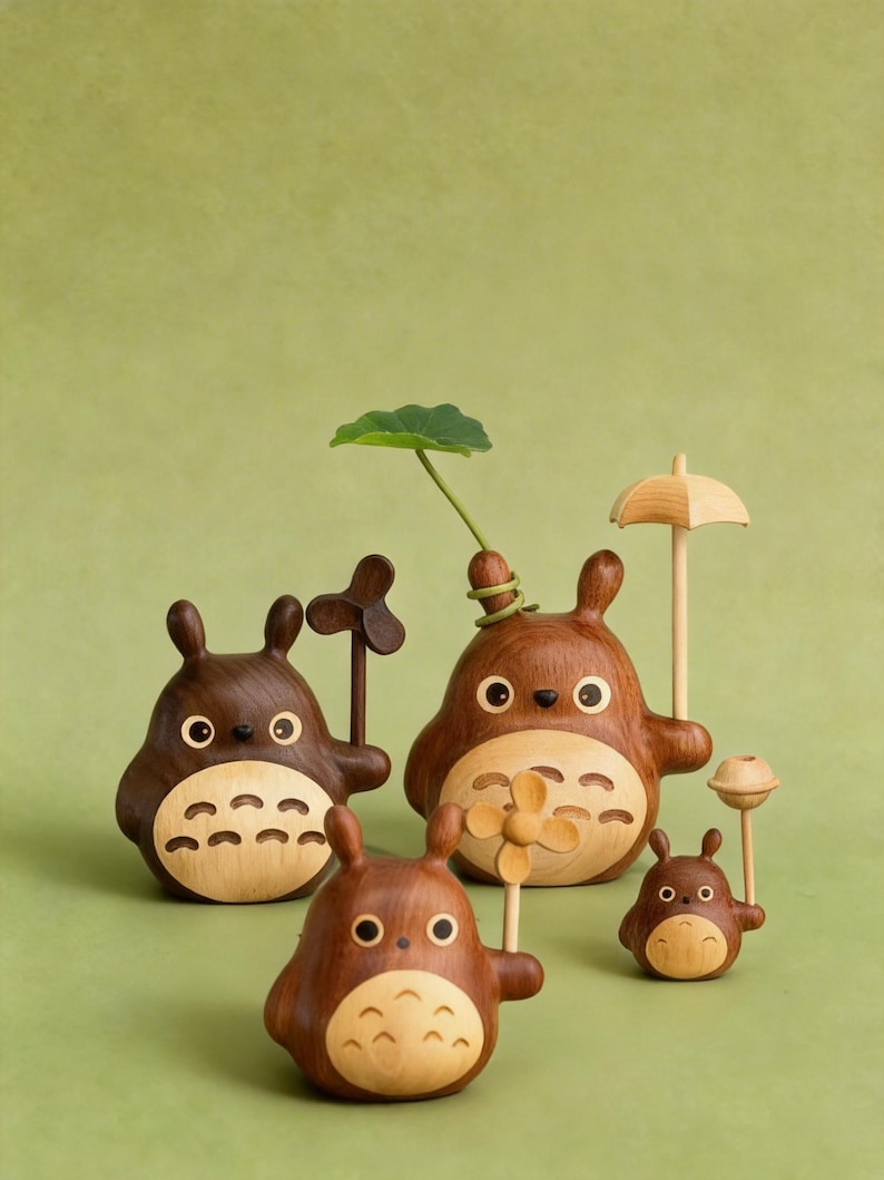 Puede incluir: Una colecci&oacute;n de figuras de madera de Totoro de varios tama&ntilde;os. Las figuras son marrones con vientres de madera clara y presentan peque&ntilde;os accesorios como una hoja, un paraguas y un abanico. El fondo es verde liso.